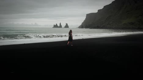 Young-beautiful-woman-in-black-dress-walking-on-black-sand-beach,-epic-aerial-ocean-scenery,-Iceland