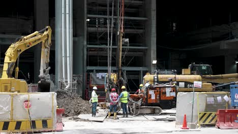 Construction-Workers-Standing-Around-At-Site-With-Heaving-Machinery-Drilling-On-Ground-In-Singapore
