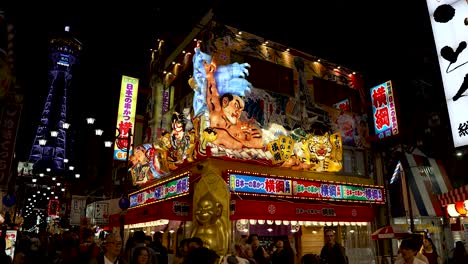 Beleuchtete,-Farbenfrohe-Ausstellung-über-Dem-Restaurant-Mit-Biliken-Statue-An-Der-Ecke-Im-Shinsekai-Viertel-Bei-Nacht-Mit-Vorbeigehenden-Menschen