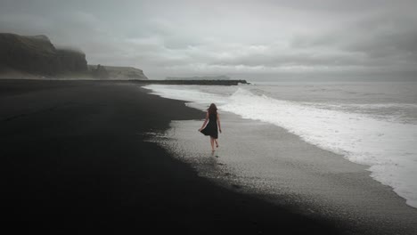 Young-beautiful-woman-in-black-dress-running-on-black-sand-beach-Iceland,-aerial-dramatic-waves-seascape