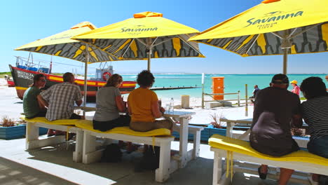 Tourists-sit-under-yellow-restaurant-umbrellas,-scenic-Struisbaai-harbour-view