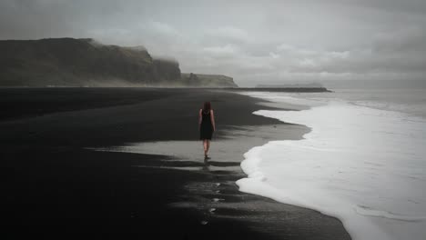 Young-beautiful-woman-in-black-dress-walking-on-black-sand-beach-Iceland,-waves-flowing-,-tracking-shot