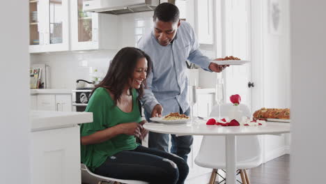 Frau-Mittleren-Alters-Sitzt-In-Der-Küche,-Während-Ihr-Partner-Ihnen-Ein-Romantisches-Essen-Serviert