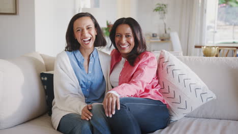 Mother-and-adult-daughter-sitting-on-sofa-together-laughing-to-camera,-close-up