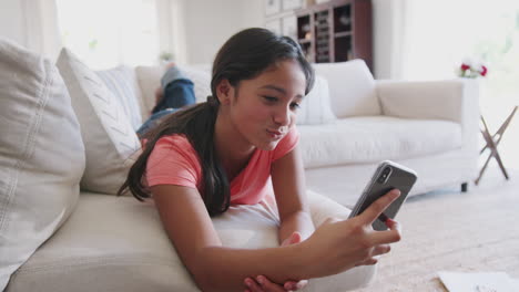 Happy-teenage-African-American-girl-lying-on-a-sofa-at-home-using-smartphone,-low-angle,-close-up