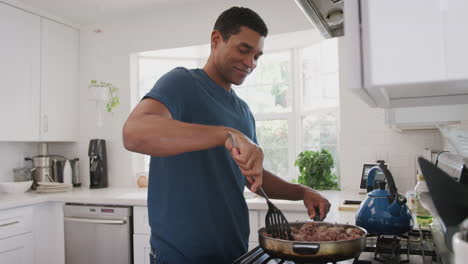 Millennial-African-American-man-standing-in-the-kitchen-cooking-in-a-pan-on-the-hob,side-view,waist-up