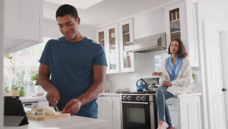 Young-African-American-man-preparing-food-in-the-kitchen,-his-partner-sitting-on-kitchen-worktop-behind-him