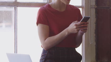 Millennial-Hispanic-woman-standing-by-window-in-a-creative-office-using-smartphone,-tilt-shot
