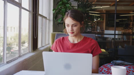 Millennial-Hispanic-female-creative-sitting-at-a-desk-using-laptop-in-an-office,-panning-shot