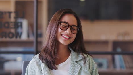 Millennial-Asian-female-creative-wearing-glasses,-sitting-in-an-office-smiling-to-camera,-close-up