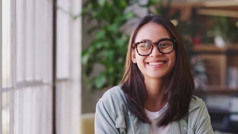 Millennial-Asian-female-creative-in-glasses-standing-in-office-turns-and-smiles-to-camera,-close-up