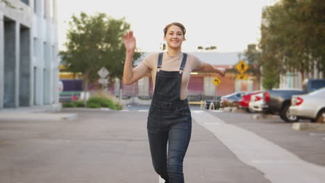 Young-Hispanic-woman-in-dungarees-running-to-camera-in-a-city-street,-full-length,-close-up