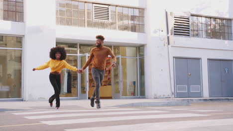Fashionable-young-black-couple-running-across-a-city-street-holding-hands,-full-length