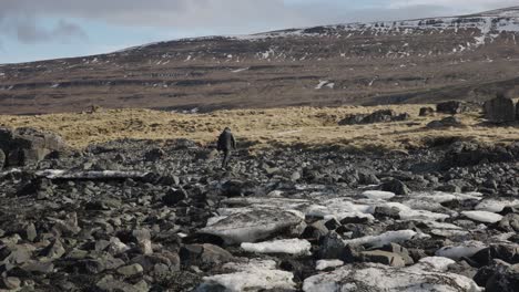 Man-walk-on-black-volcanic-rocks-with-huge-mountain-in-front,-Iceland