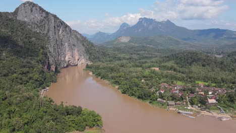 Luftaufnahme-Des-Mekong-Flusses-Neben-Der-örtlichen-Dorfstadt-Und-Hoch-Aufragenden-Klippen-In-Luang-Prabang