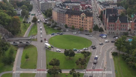 Drone-footage-of-traffic-driving-in-roundabout-in-Roslagstull,-Stockholm,-Sweden-on-overcast-day-with-lush-green-grass-and-trees-at-start-of-fall