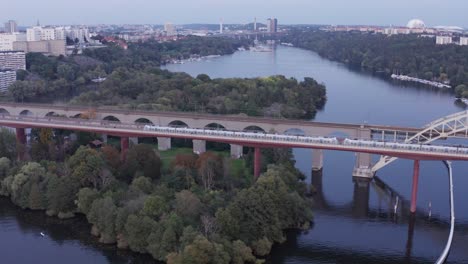 Bridges-with-public-transportation-train-going-by-over-water-and-with-trees-in-Stockholm,-Sweden-during-sunset