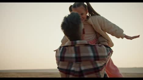 A-happy-middle-aged-brunette-man-with-gray-hair-in-a-plaid-shirt-lifts-his-little-daughter-a-blonde-girl-in-a-white-jacket-and-spins-with-her-during-his-vacation-on-a-hike-on-a-deserted-seashore-in-a-summer-evening