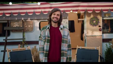 Portrait-of-a-Happy-brunette-guy-with-stubble-in-a-Green-checkered-shirt-and-a-red-T-shirt-who-stands-near-his-decorated-trailer-during-his-picnic-outside-the-city-at-a-camp-in-the-summer