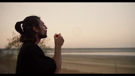 A-man-with-long-brunette-hair-and-stubble-blows-soap-bubbles-during-his-vacation-outside-the-city-on-a-summer-evening.-A-mysterious-man-blows-soap-bubbles-while-relaxing-near-a-pond-on-a-deserted-shore-in-the-summer-evening