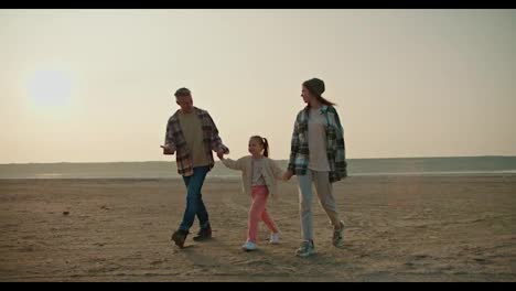 A-happy-brunette-man-in-a-checkered-shirt-walks-hand-in-hand-with-his-little-daughter,-a-blonde-girl-in-a-white-jacket-and-pink-pants,-and-his-wife,-a-brunette-girl-in-a-checkered-green-shirt,-during-his-vacation-outside-the-city-in-the-summer-evening