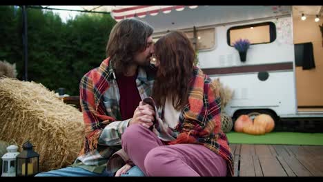 Close-up-shot-of-a-happy-couple,-brunette-guy-in-a-green-checkered-shirt-sits-wrapped-in-a-red-checkered-blanket-with-his-brunette-girlfriend-near-their-trailer-in-a-camp-outside-the-city-during-a-picnic-in-the-summer