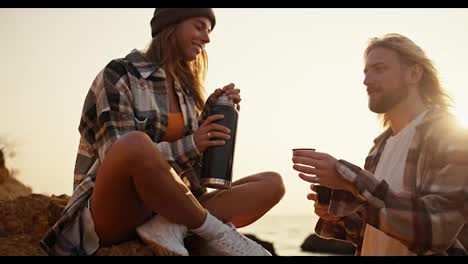 happy-blonde-girl-in-a-checkered-shirt-pours-hot-tea-from-a-black-thermos-to-her-blonde-boyfriend-with-a-beard-into-a-special-mug,-sitting-on-a-large-stone-on-a-rocky-shore-near-the-sea-in-the-morning