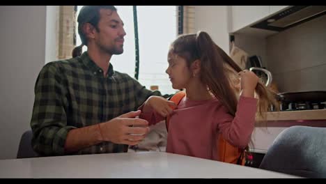 A-happy-brunette-man-in-a-green-checkered-shirt-helps-his-little-daughter,-a-brunette-girl-in-a-pink-dress,-put-an-orange-backpack-on-her-back-and-in-the-kitchen-in-a-modern-apartment