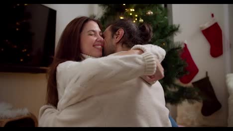 Happy-couple-guy-and-girl-sitting-opposite-each-other-and-hugging-in-white-sweaters-near-the-Christmas-tree-in-a-cozy-room-in-winter.-The-magic-of-Christmas-warmth-and-comfort-as-well-as-the-family-atmosphere-in-winter