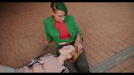 Top-view-of-two-lesbian-girls-during-their-date-communicating-with-each-other-against-the-backdrop-of-paving-stones.-A-blonde-girl-with-a-short-haircut-in-a-plaid-shirt-lies-on-the-lap-of-her-girlfriend-with-a-green-haircut-and-a-green-shirt-on-her-date-LGBT-couple