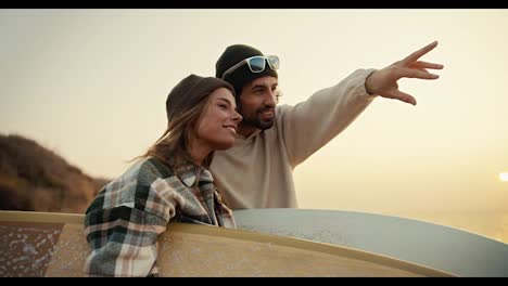 A-brunette-man-in-a-black-hat-and-white-top-points-somewhere-in-the-direction-of-his-blonde-girlfriend-in-a-plaid-shirt-while-they-stand-on-the-seashore-at-dawn-in-the-morning-and-hold-surfboards-in-their-hands