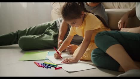 Close-up-shot-of-a-little-happy-brunette-girl-in-a-yellow-dress-sitting-on-the-floor-near-her-parents-and-drawing-on-paper-using-multi-colored-felt-tip-pens-in-a-modern-apartment