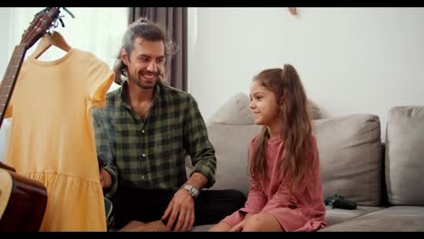 A-lonely-father,-a-brunette-man-in-a-green-checkered-shirt,-communicates-with-his-daughter-and-steams-a-yellow-dress-using-a-steamer-while-sitting-on-a-gray-sofa-in-a-modern-apartment