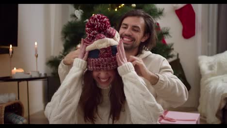 Portrait-of-a-young-brunette-guy-puts-a-woolen-New-Year's-hat-on-his-girlfriend's-head-in-a-White-sweater-in-a-room-with-a-New-Year's-tree-and-a-cozy-atmosphere-in-a-winter-evening