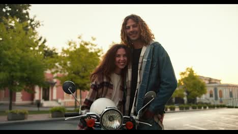 Portrait-of-a-happy-guy-and-girl-with-curly-hair-in-plaid-shirts,-a-girl-sits-on-a-moped,-holds-a-white-helmet-in-her-hands-and-looks-at-her-boyfriend-in-a-denim-jacket-on-the-street-near-the-trees-on-a-wide-road-in-summer