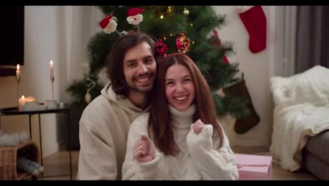 portrait-of-a-happy-brunette-guy-who-sits-with-his-brunette-girlfriend-in-a-white-sweater-who-moves-her-hands-back-and-forth-to-the-beat-of-the-music-near-the-New-Year-tree-in-a-room-decorated-for-the-New-Year-in-winter