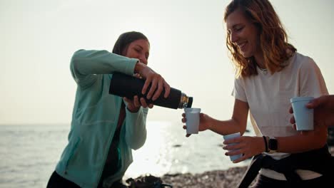 A-girl-in-a-blue-jacket-pours-tea-from-a-thermos-to-a-blonde-girl-with-a-white-T-shirt-during-their-rest-and-relaxation-during-rock-climbing.-A-group-of-rock-climbers-drink-tea-during-their-break-between-rock-climbs