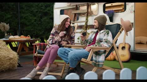 A-happy-brunette-girl-in-a-hat-and-a-plaid-shirt-together-with-her-brunette-boyfriend-in-a-green-plaid-shirt-are-drinking-tea,-sitting-on-chairs-and-talking-near-a-trailer-during-their-picnic-in-a-camp-outside-the-city-in-the-summer