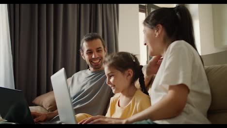 Familie-Sitzt-Auf-Dem-Sofa-Und-Schaut-Sich-Etwas-Auf-Einem-Grauen-Laptop-An.-Ein-Brünettes-Mädchen-In-Einem-Weißen-T-Shirt,-Ihre-Tochter,-Ein-Mädchen-In-Einem-Gelben-Kleid-Und-Ein-Brünetter-Mann-In-Einem-Grauen-T-Shirt-Sitzen-Zusammen-Auf-Einem-Hellbraunen-Sofa-Und-Schauen-Auf-Den-Bildschirm-Eines-Grauen-Laptops-In-Einer-Modernen-Wohnung