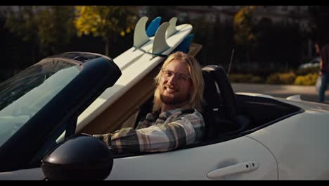 Portrait-of-a-happy-blond-guy-in-glasses-with-a-beard-in-a-checkered-shirt-who-sits-in-a-White-convertible-and-to-the-right-of-him-there-are-two-surfboards-in-the-city