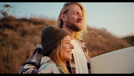 Close-up-shot-of-a-happy-blond-guy-with-a-beard-in-a-plaid-shirt-hugs-his-blonde-girlfriend-in-a-hat-in-a-plaid-shirt-and-holds-a-surfboard-near-the-strawberry-seashore-at-Sunrise-in-the-morning-in-summer