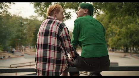 Rear-view-of-two-LGBT-girls-sitting-on-a-fence-in-a-skate-park-talking-and-stroking-each-other.-A-blonde-girl-with-a-short-haircut-VK-what-are-you-wearing-a-shirt-and-a-girl-with-green-hair-with-a-short-haircut-in-a-Green-shirt-during-her-LGBT-date-in-the-park