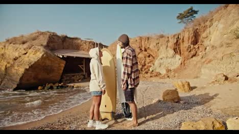 A-brunette-man-in-a-checkered-shirt-stands-with-his-blonde-girlfriend-in-a-white-sweatshirt,-looks-at-her-and-communicates,-they-hold-surfboards-in-their-hands-while-standing-on-a-sandy-rocky-beach-near-the-sea-in-the-morning