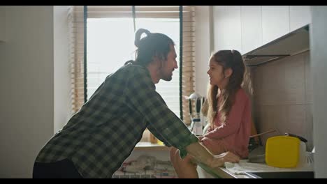 Close-up-shot-of-a-brunette-man-in-a-green-checkered-shirt-leaning-on-the-table-and-talking-with-his-little-brunette-daughter-in-a-pink-dress-in-a-modern-white-kitchen