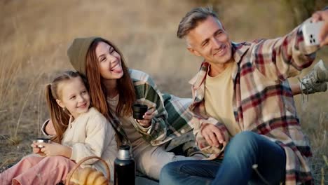 A-happy-middle-aged-man,-together-with-his-wife,-a-brunette-girl-in-a-Green-checkered-shirt,-and-a-little-daughter,-take-a-selfie-using-a-white-phone-during-their-picnic-and-vacation-outside-the-city-in-the-summer