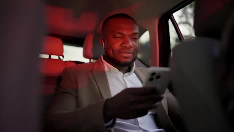 A-confident-man-with-Black-skin-in-a-brown-suit-with-a-short-haircut-and-beard-rides-in-the-interior-of-a-modern-car-and-works-on-a-white-smartphone