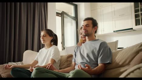 A-brunette-girl-in-a-white-T-shirt-together-with-her-husband,-a-brunette-man-in-a-gray-T-shirt,-meditates-while-sitting-on-light-brown-daughter-girl,-a-little-brunette-girl-in-a-yellow-dress-interferes-with-them-and-has-fun-in-a-modern-studio-apartment