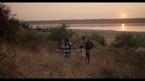 Rear-view-of-a-brunette-girl-in-a-green-checkered-shirt-along-with-her-brunette-husband-and-little-daughter-walking-down-the-hill-after-their-picnic-outside-the-city-in-the-summer-evening