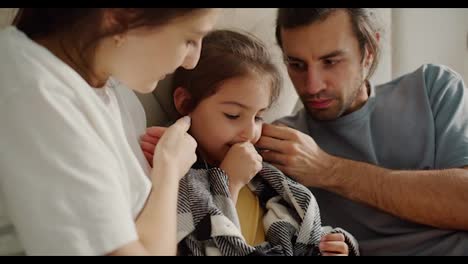 Two-parents,-a-brunette-man-in-a-gray-T-shirt-and-his-wife,-a-brunette-girl-in-a-white-T-shirt,-are-caring-for-and-stroking-their-sick-daughter-who-is-sitting-in-a-blanket-on-the-sofa