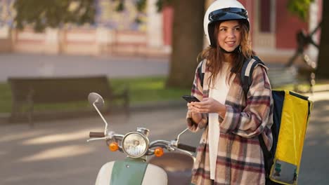 A-brunette-courier-girl-in-a-checkered-shirt-in-a-White-helmet-for-a-moped-stands-near-her-moped-with-a-large-yellow-bag,-looks-at-her-phone-at-the-navigator-and-looks-around-the-area-to-understand-where-to-deliver-food